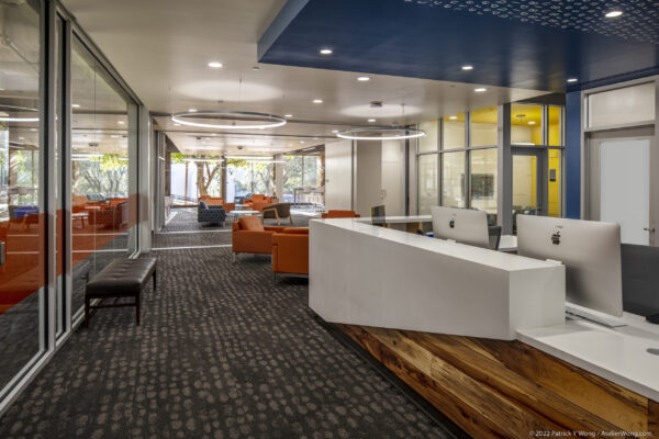 Room with gray carpet and white couches. There are large glasses panels for each office and orange couches sprinkled throughout the room.