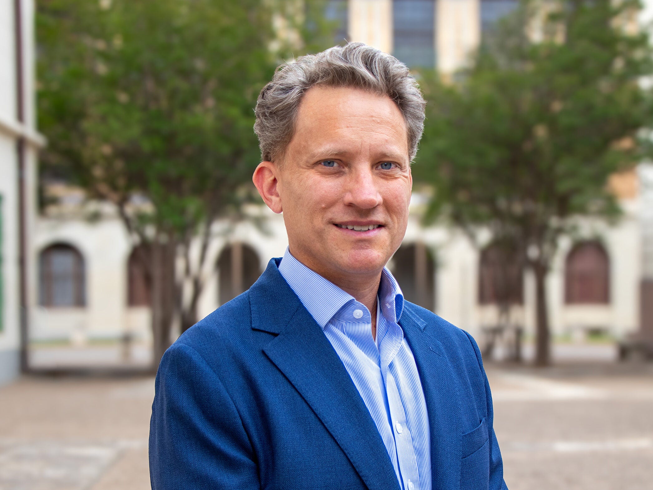 A professionally dressed man in a blue blazer stands outdoors in a courtyard with trees and historic buildings blurred in the background