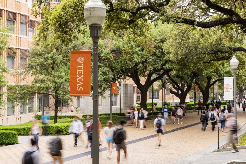 Photo of a tree-lined university campus walkway on a sunny day, with students walking in both directions along a stone path. An orange banner reading 'TEXAS' hangs from a lamppost in the foreground, and brick buildings with white columns are visible on the left through the trees.